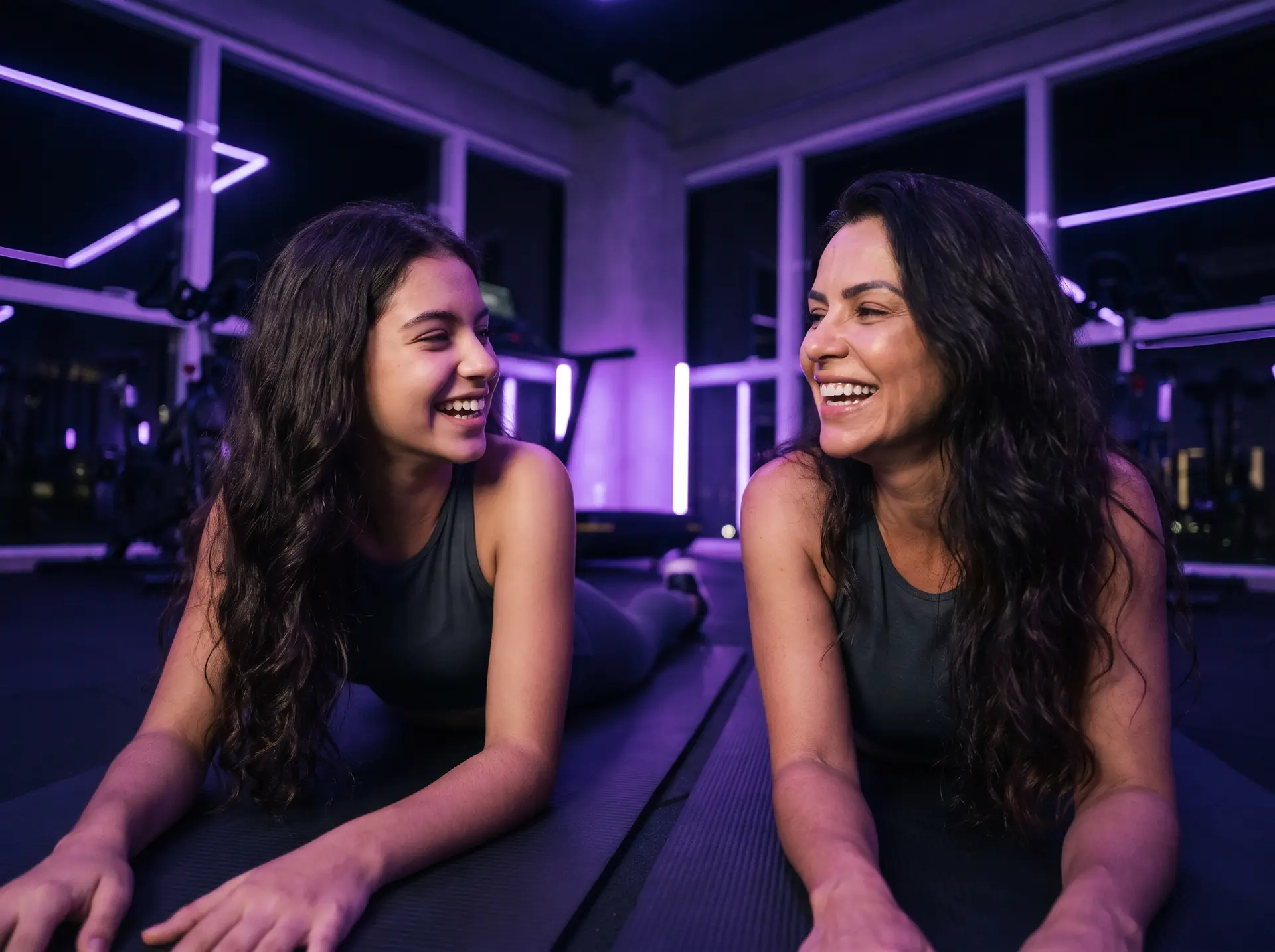 Mãe e filha rindo juntas sobre colchonetes em uma academia.
