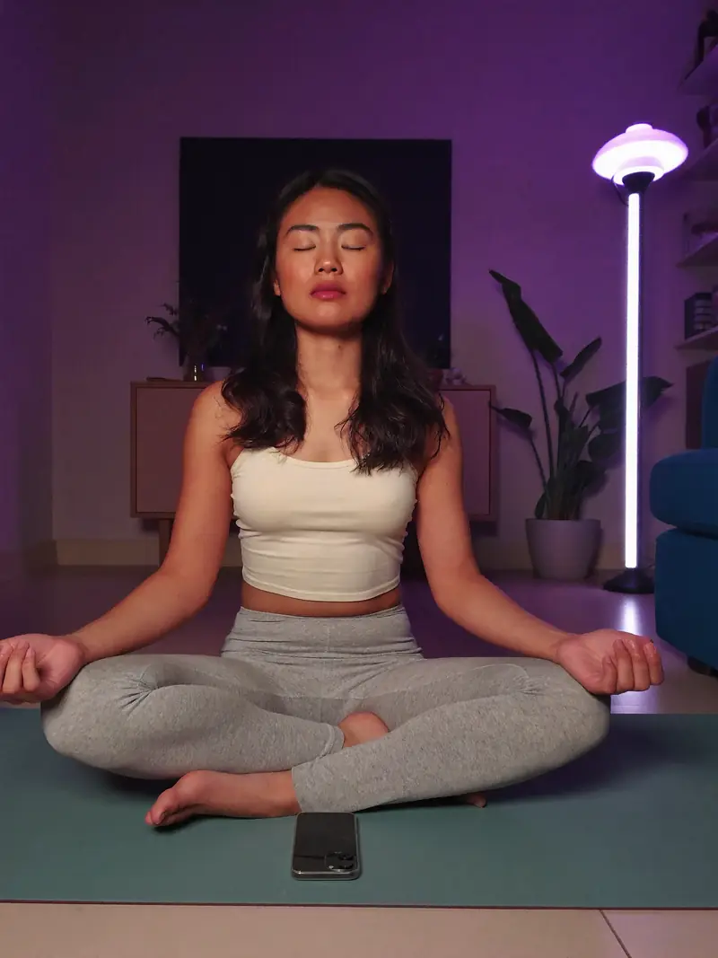 Mulher meditando sobre um tapete, com o celular ao lado.