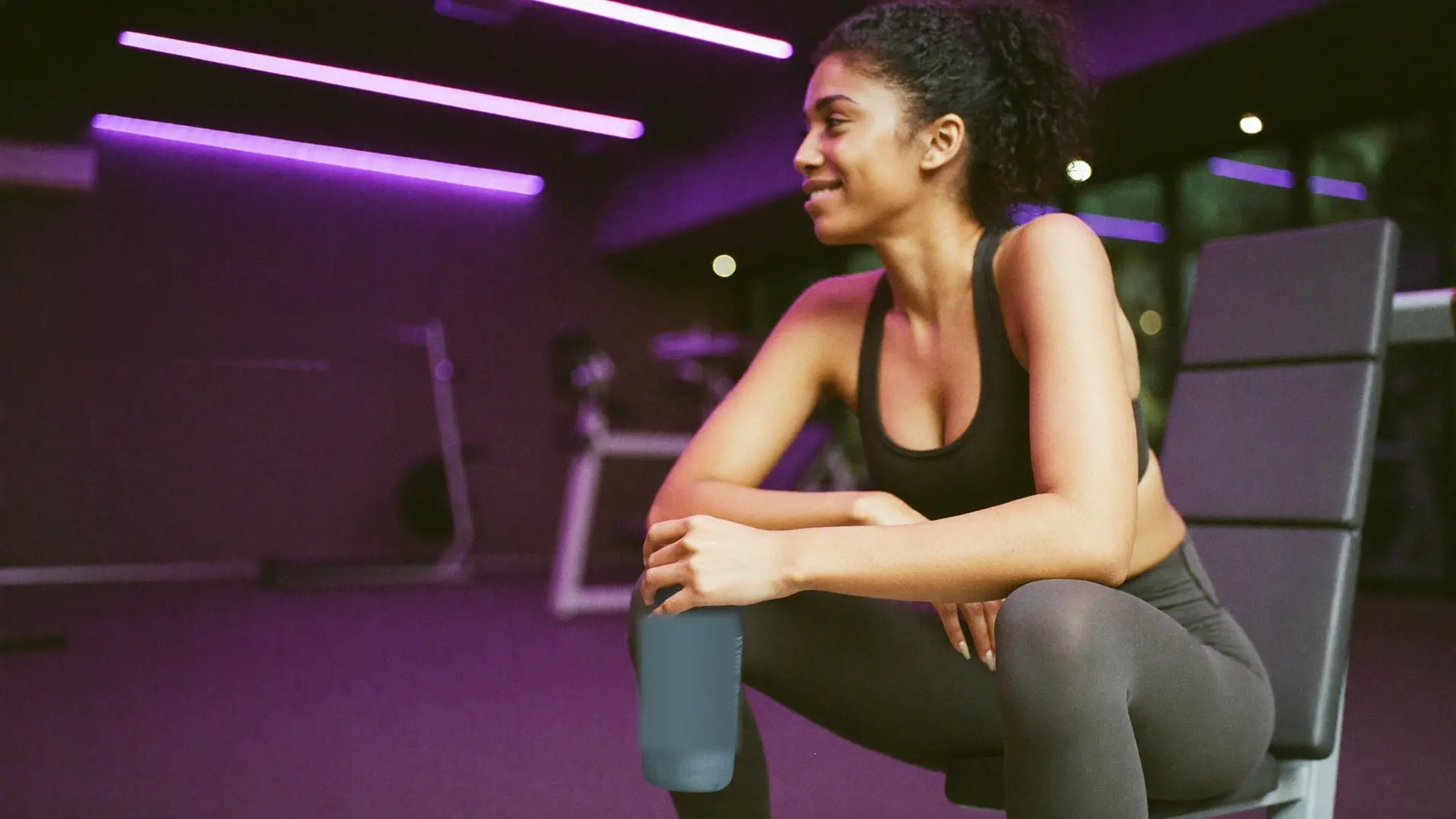 Mulher sorrindo sentada em um banco de academia, com luzes violeta ao fundo.