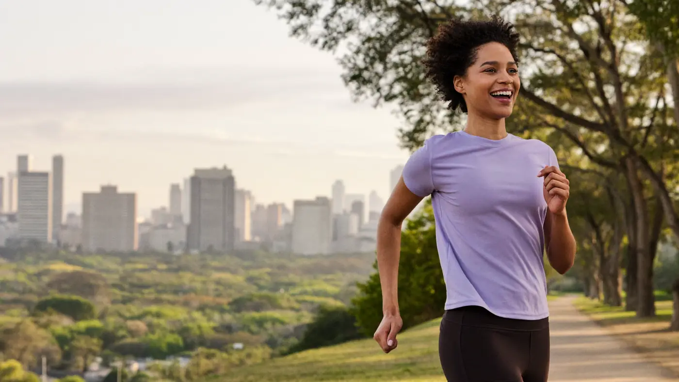 Mulher correndo sorridente em um parque, com prédios ao fundo.